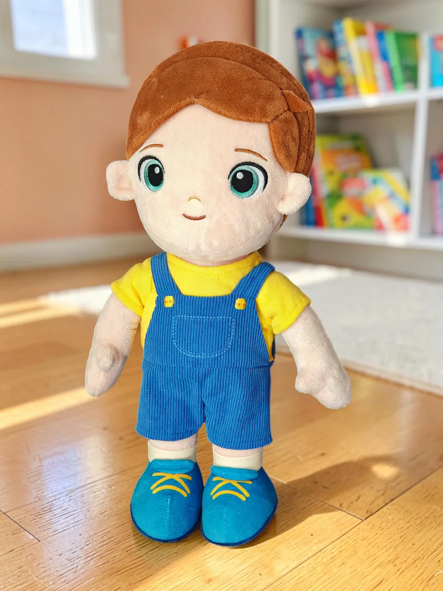 Levi, the Playnitive boy doll, standing on a wooden floor in a child's room: brown hair, a yellow shirt and blue corduroy dungarees